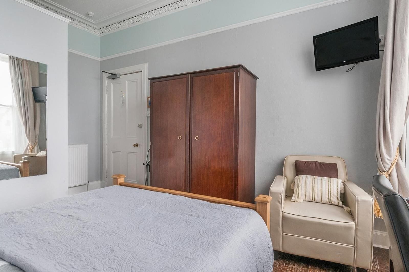 Un dormitorio con una cama, un armario de madera, una silla y un televisor montado en la pared.