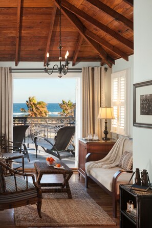 Une pièce chaleureuse avec un plafond en bois, un balcon offrant une vue sur l’océan, des meubles en osier et une table ornée d’oranges.