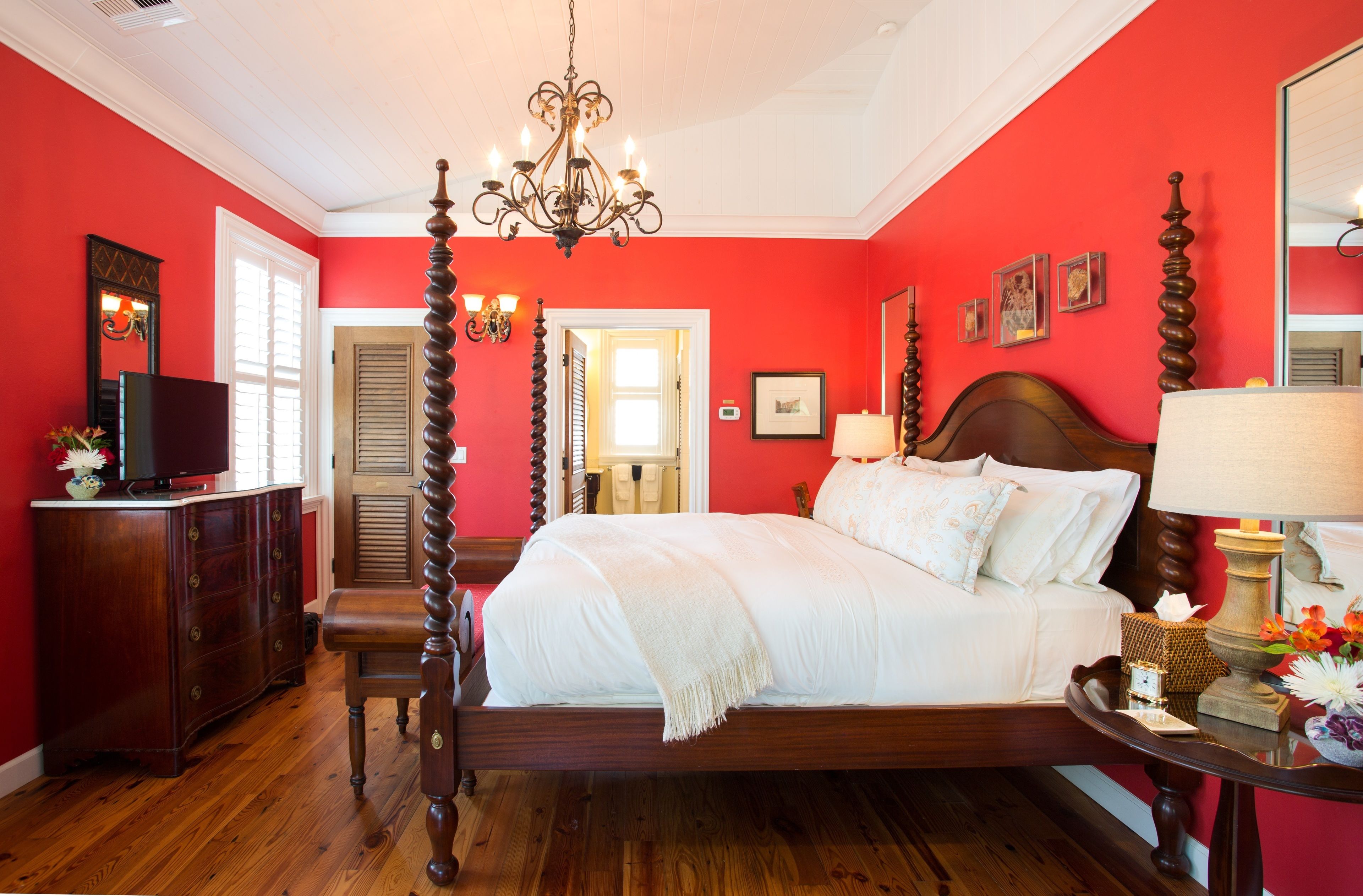 A bedroom with a large bed, wooden headboard, and a chandelier. There is a dresser with a mirror and a television on top.