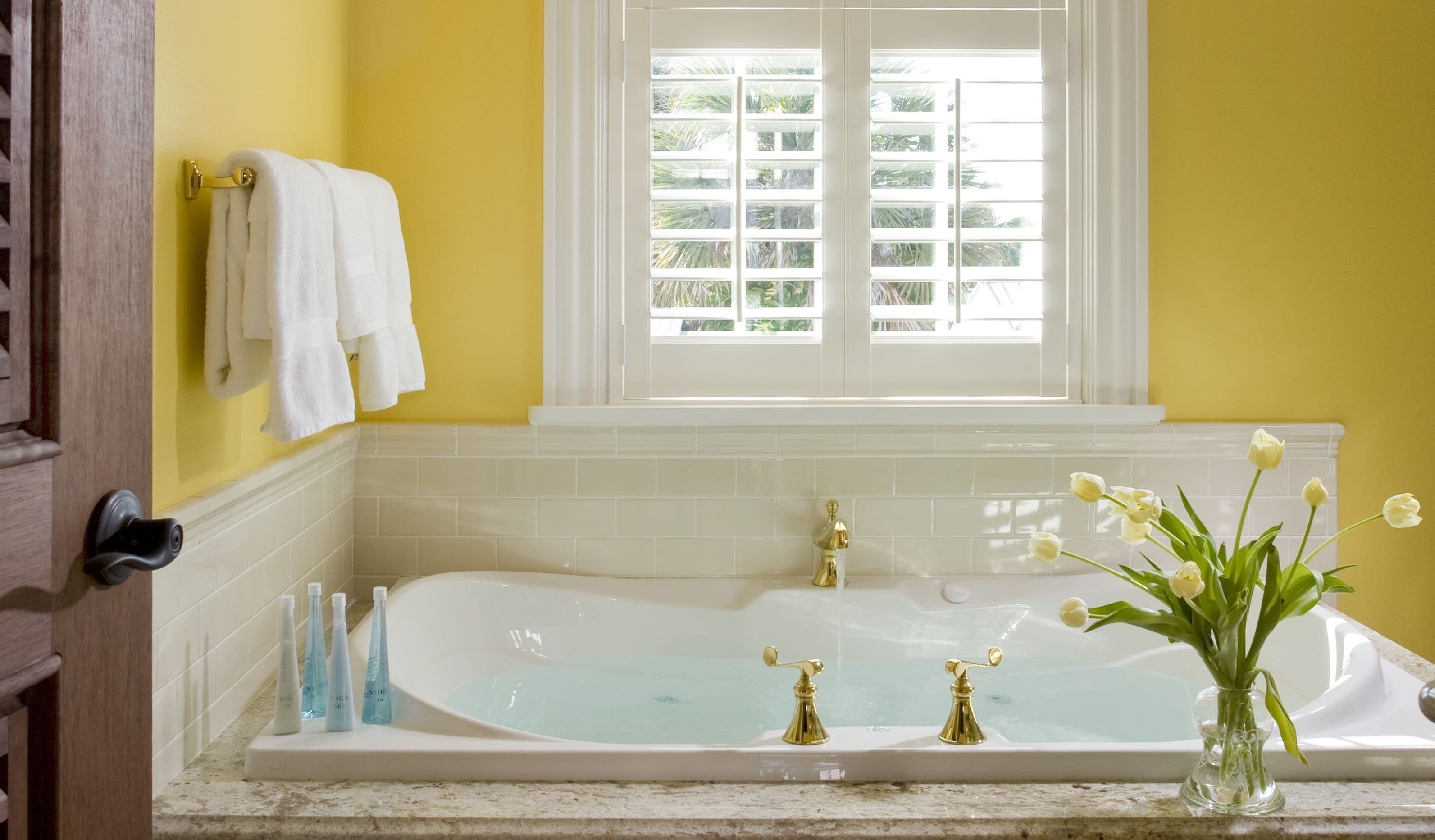 Un baño con bañera, ventana con vista a palmeras y un florero con flores en la encimera.