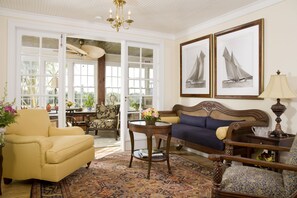 A well-lit living room with a sofa, armchairs, a coffee table, and a chandelier. There are two framed pictures on the wall and a window with a view of the outdoors.