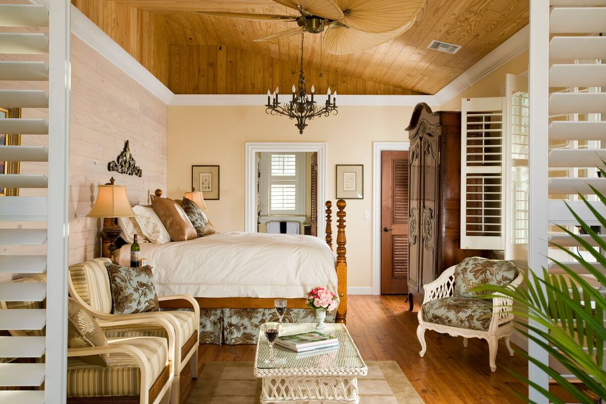 A bedroom with a large bed, two chairs, a small table with a book and a glass, and a chandelier.