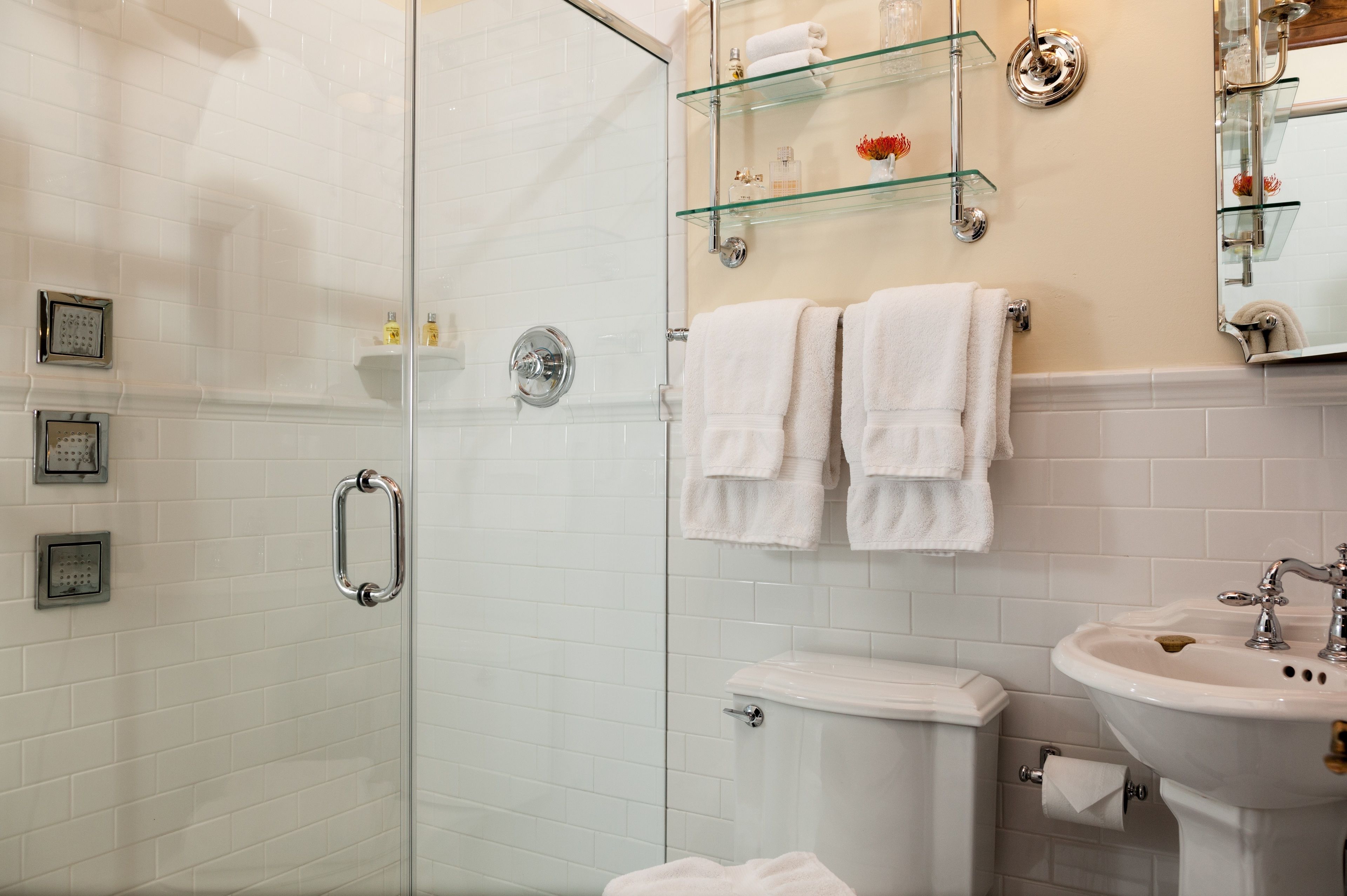 Un baño moderno con cabina de ducha de vidrio, inodoro, lavamanos y estantes de vidrio con artículos de aseo.