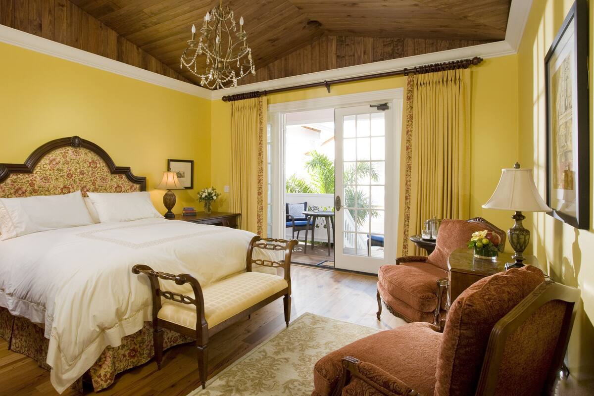 A bedroom with a large bed, two chairs, a chandelier, and a view of the outdoors.