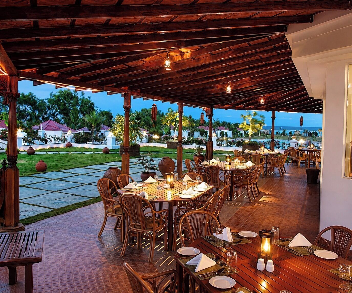 Un espace de restauration en plein air, avec des tables et des chaises en bois, agrémenté d’un jardin paysager en arrière-plan et offrant une vue sur la mer.