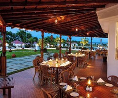 Un'area pranzo all'aperto con tavoli e sedie in legno, con sullo sfondo un giardino curato e vista sul mare.
