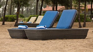 Une rangée de fauteuils de plage en osier, dotés de coussins bleus, sur une plage de sable, entourée d’arbres et, au fond, d’un bâtiment avec un toit rouge.