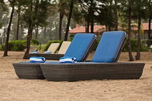 A row of wicker lounge chairs with blue cushions on a sandy beach, surrounded by trees and a building with a red roof in the background. - Chariot Beach Resort (Tirukalukundram)