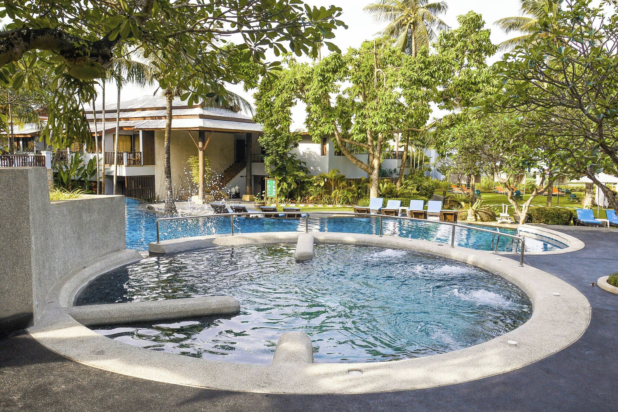 Una piscina circular con fuente, rodeada de sillones de descanso y palmeras.