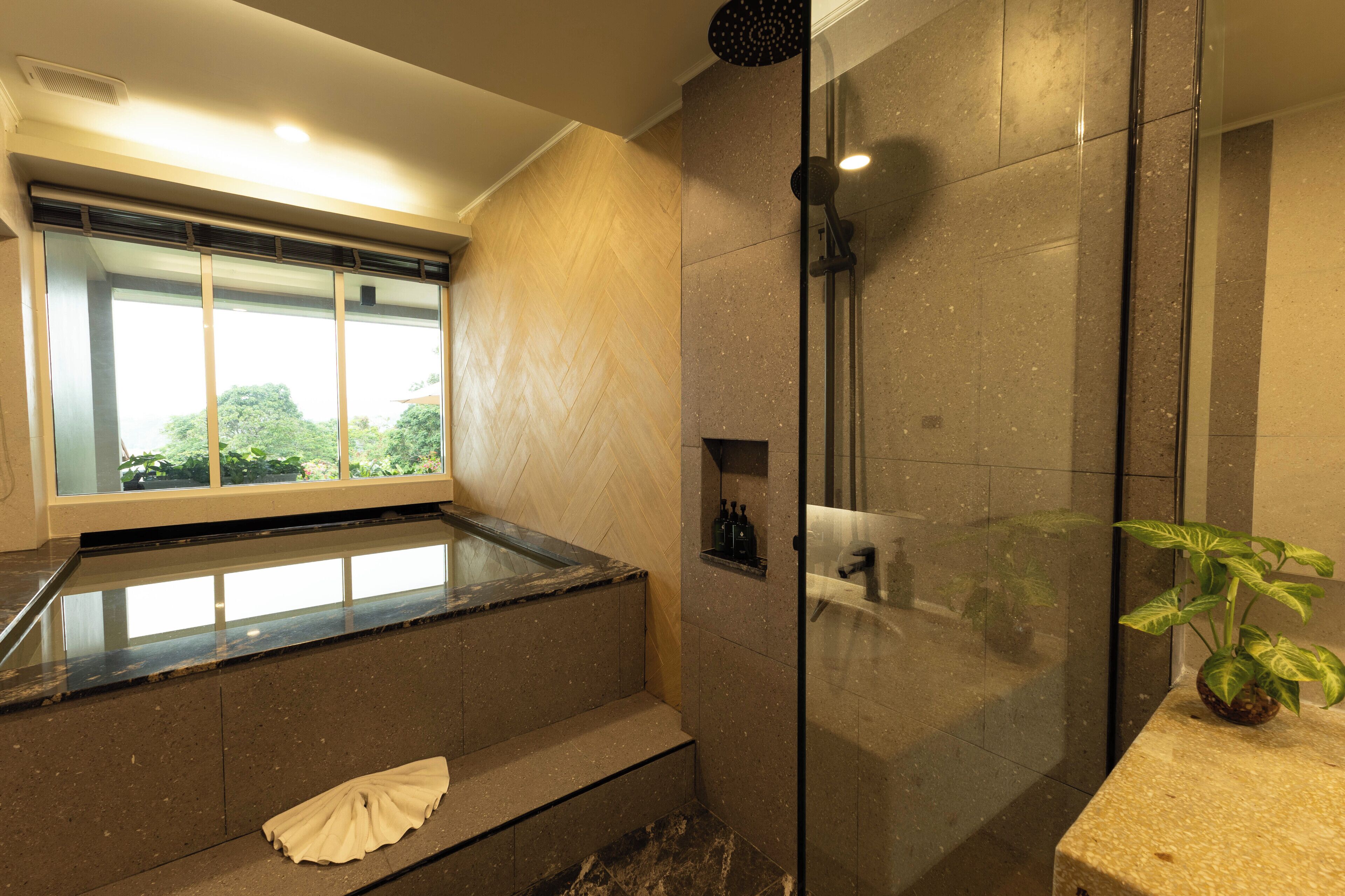 A modern bathroom with a glass shower enclosure, a bathtub, and a window offering a view of greenery outside.