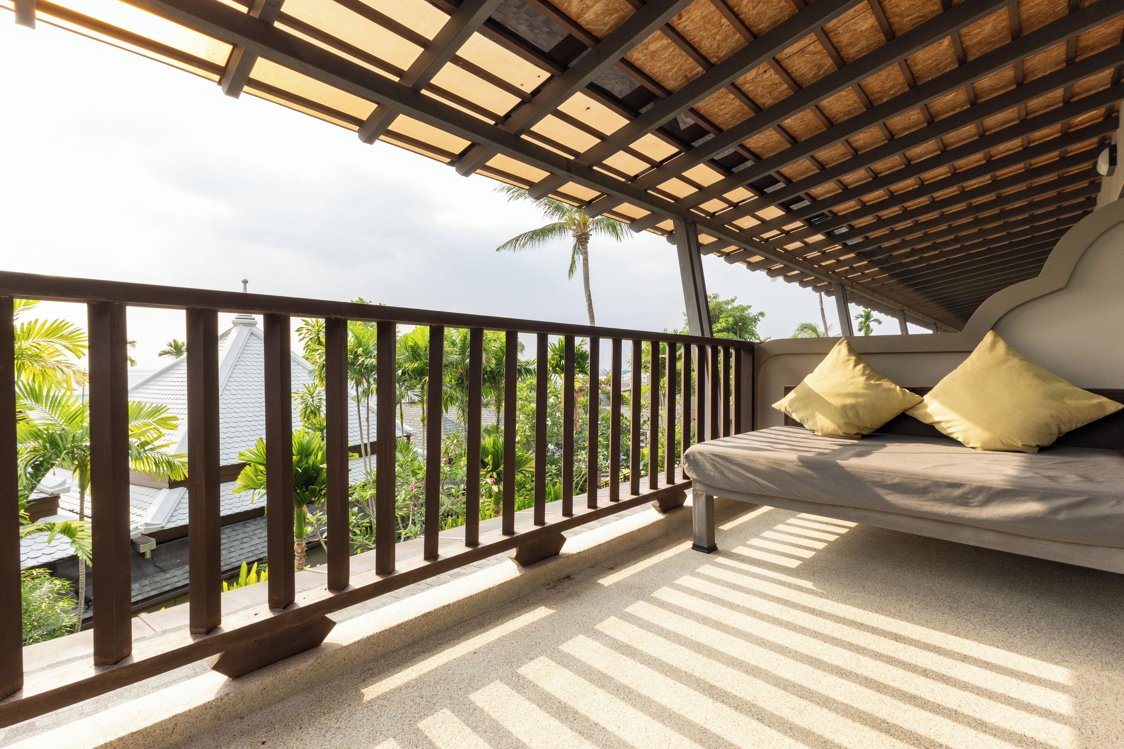 Ein Balkon mit einem Reetdach, einer Couch mit Kissen und Blick auf Palmen und Gebäude.