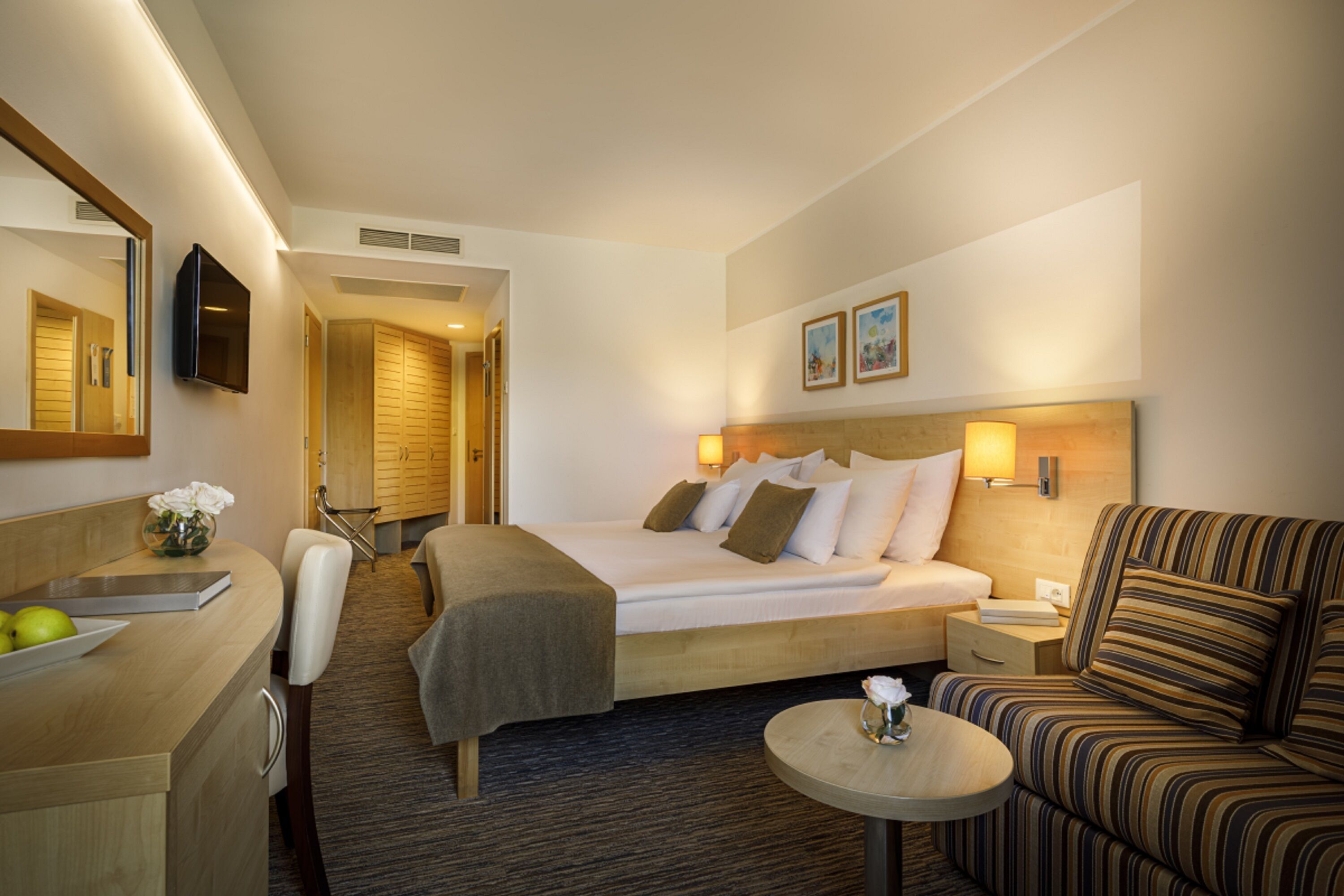 Une chambre d’hôtel avec un lit, un canapé, un bureau agrémenté d’un vase de fleurs, un miroir et un téléviseur fixé au mur.