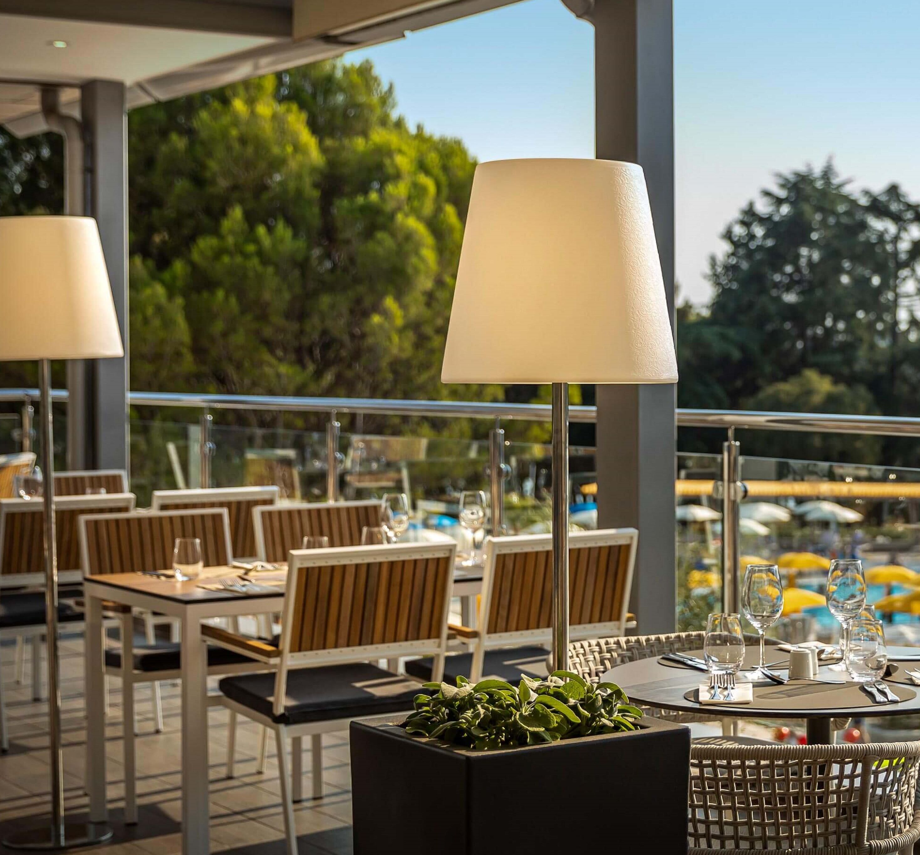 An outdoor dining area with tables and chairs, umbrellas, and a view of trees and water.