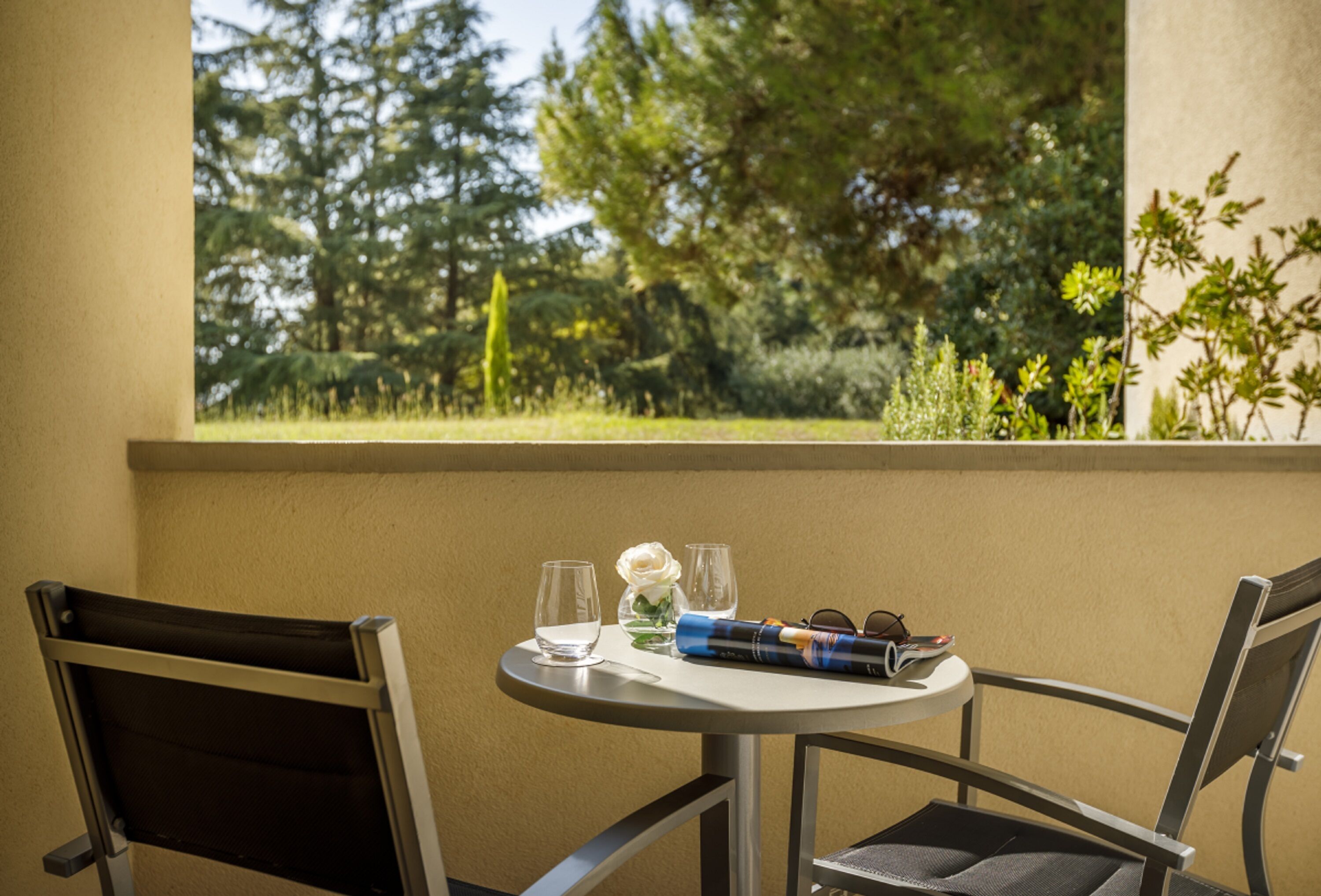 Une petite table ronde avec deux chaises, un vase contenant une fleur blanche, deux verres et un magazine ou une brochure enroulés.