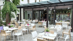 Un café al aire libre con mesas y sillas, rodeado por puertas y ventanas de vidrio.