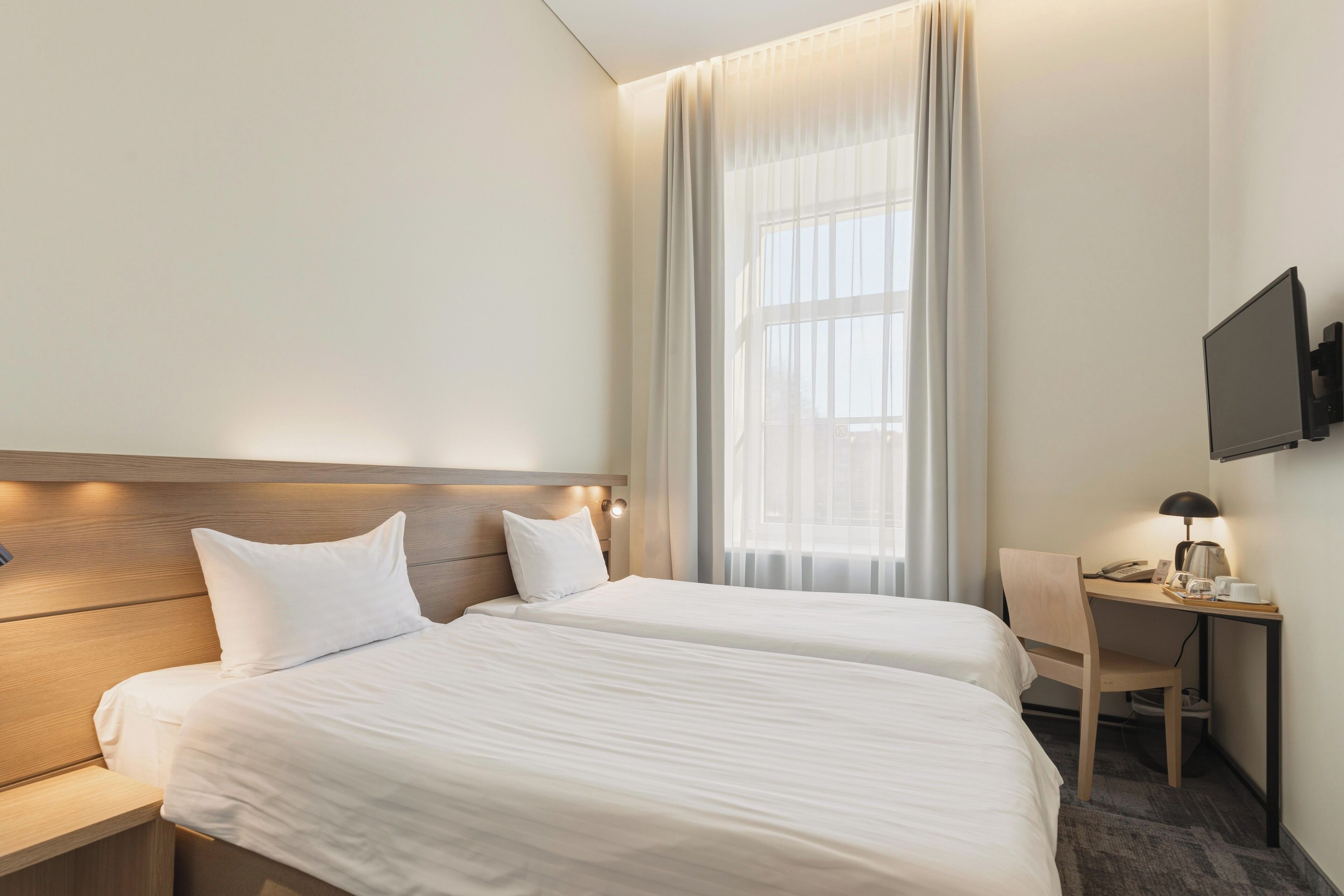 A hotel room with two beds, a desk, a chair, a TV, and a window with curtains.