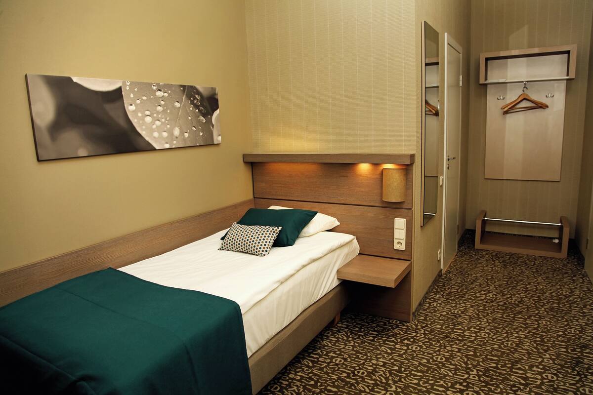 Habitación de hotel con una cama individual, una mesita de noche, un espejo, un perchero en la pared y una alfombra estampada.