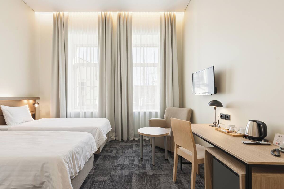 Habitación de hotel con dos camas, un escritorio con silla, televisor y ventana con cortinas.