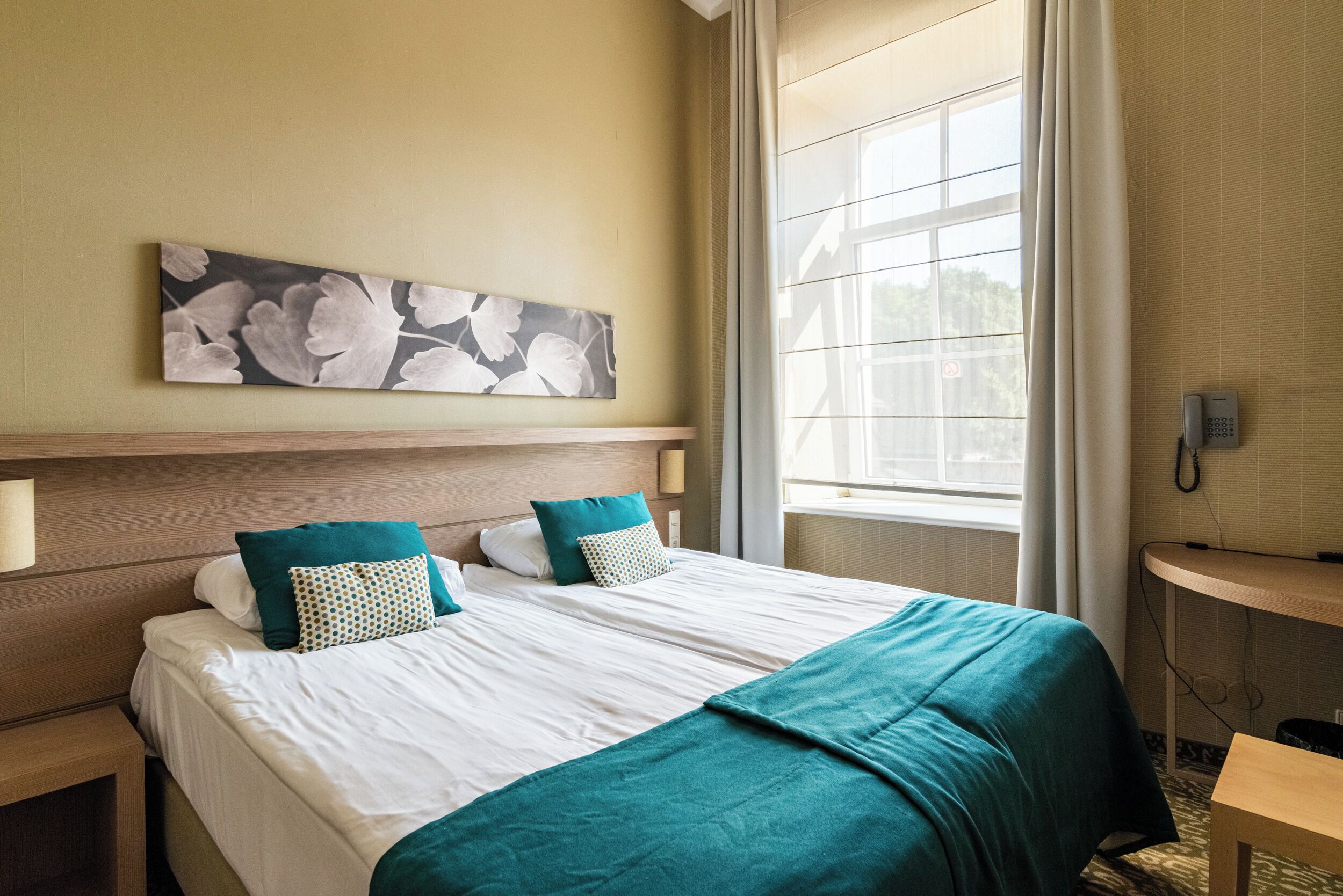 A hotel room with a bed, a bedside table, a wall-mounted phone, and a window with curtains.