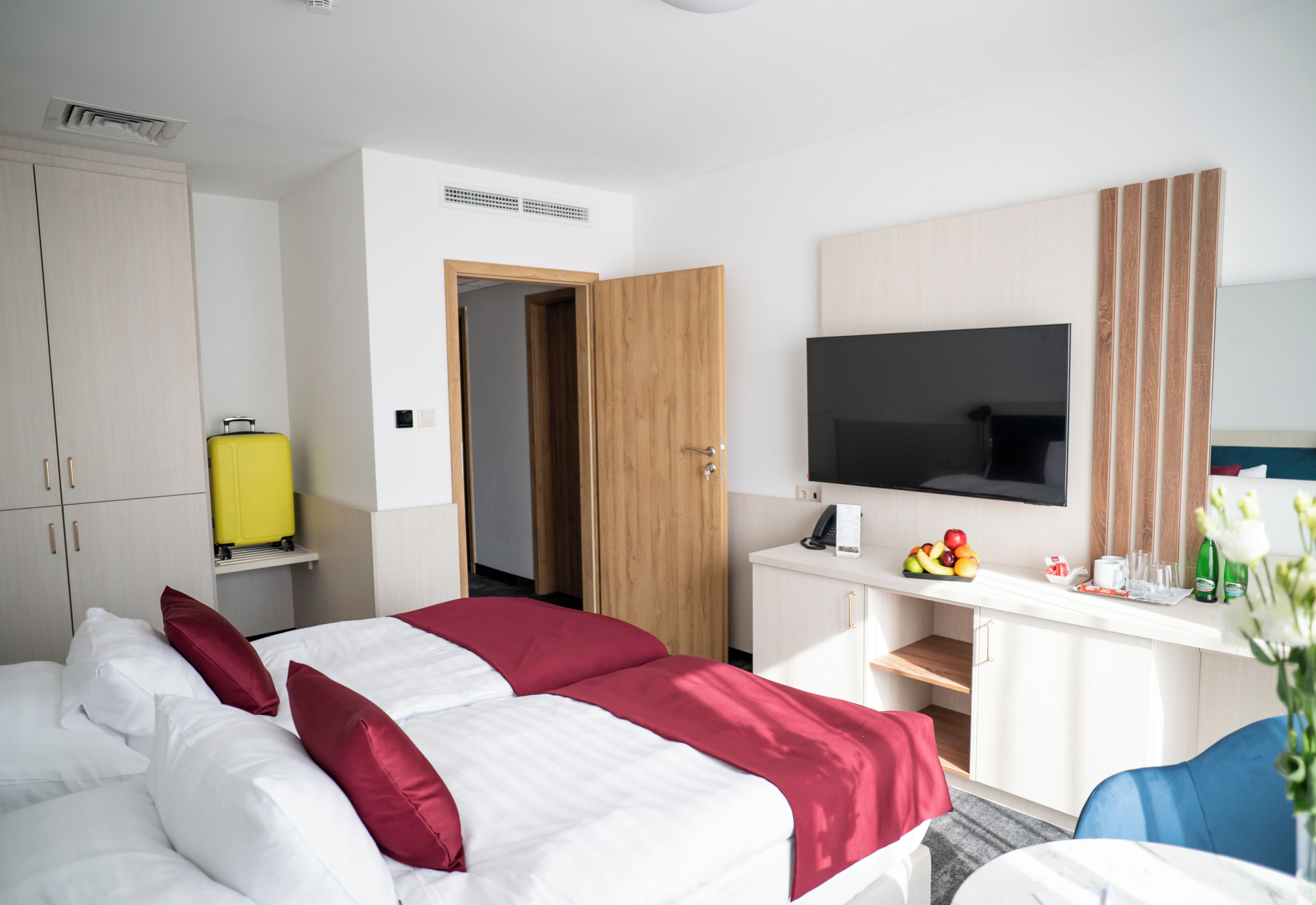 A hotel room with a bed, a desk with a TV, a wardrobe, and a small kitchenette area.
