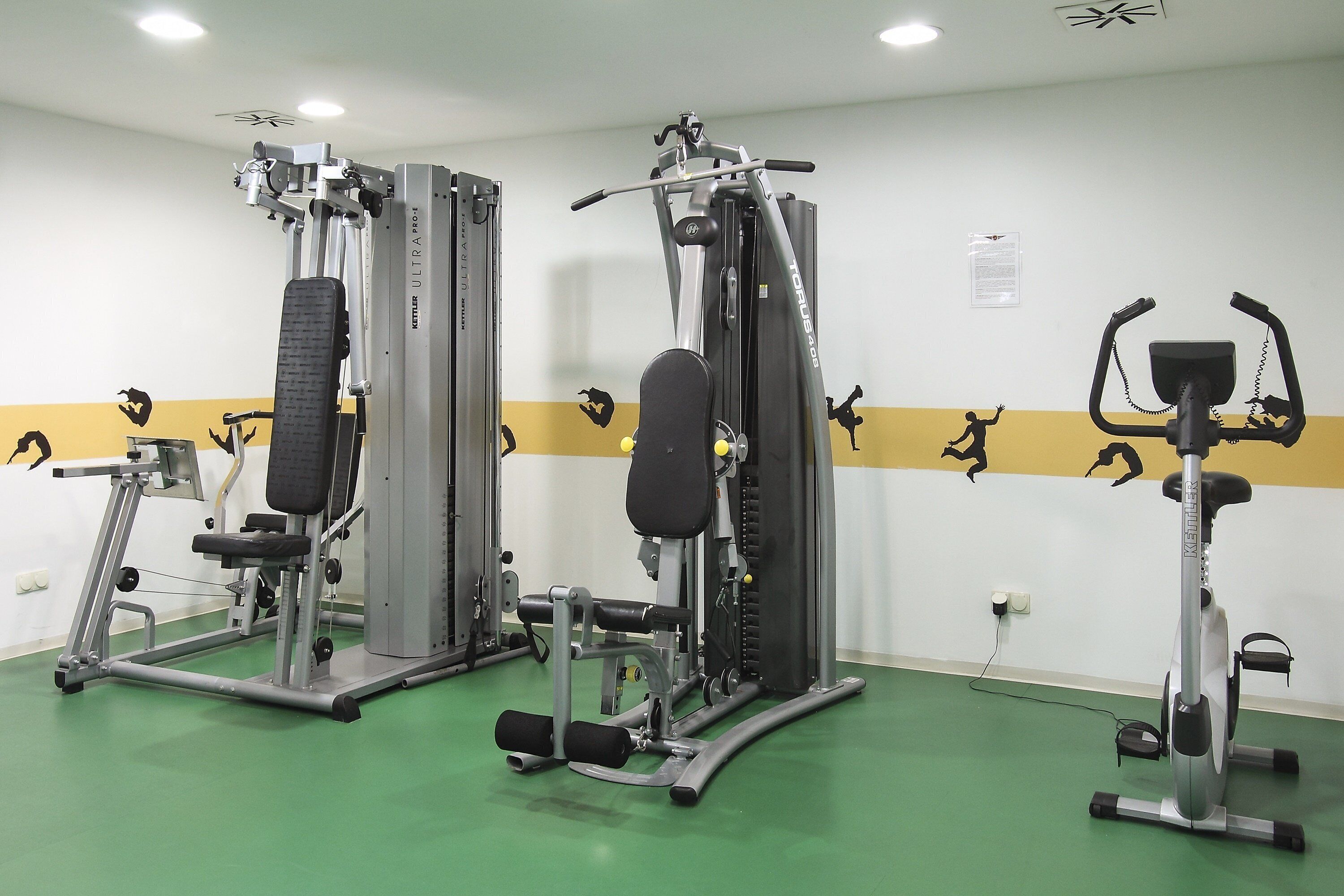 Ein Fitnessstudio mit verschiedenen Trainingsgeräten, darunter eine Beinpresse, eine Brustpresse und ein Crosstrainer.