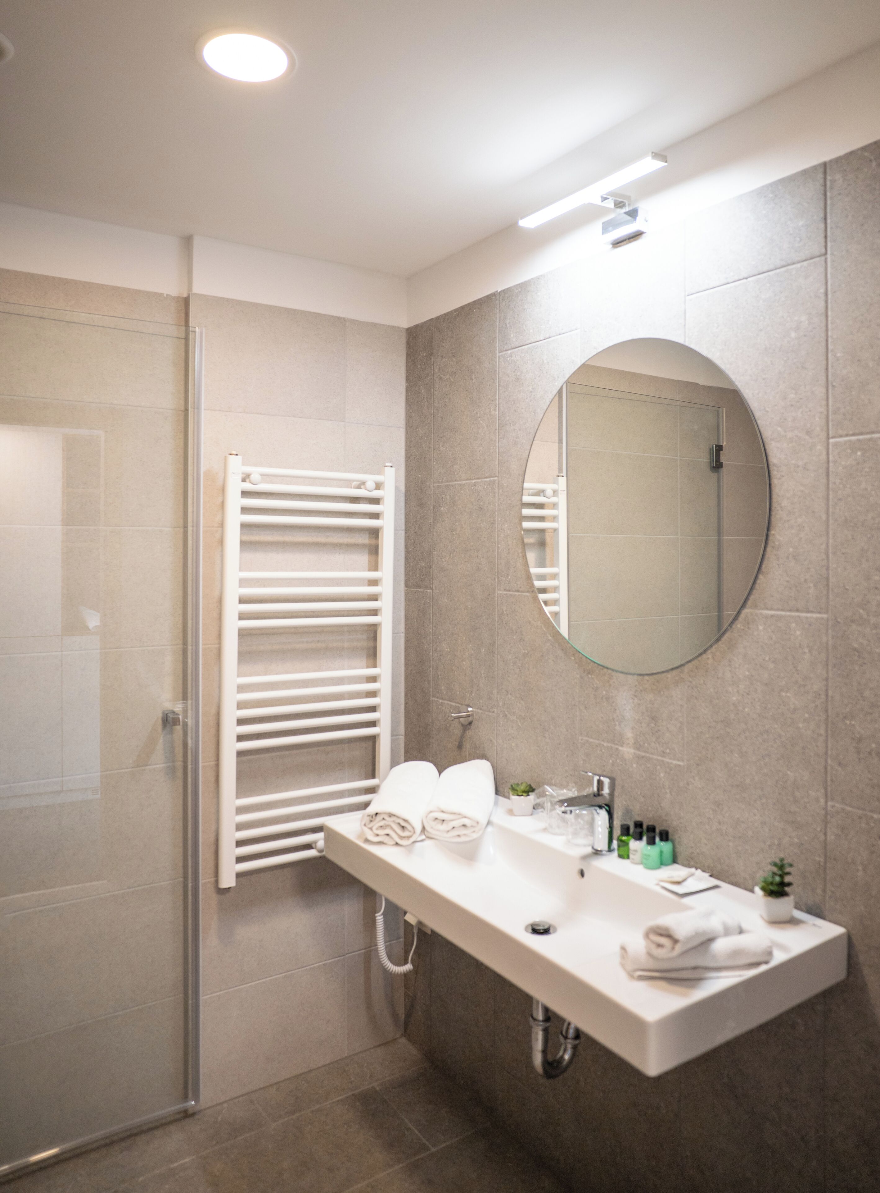 Une salle de bain moderne avec une douche à parois de verre, un porte-serviettes blanc, un grand miroir et un lavabo blanc agrémenté d’articles de toilette.