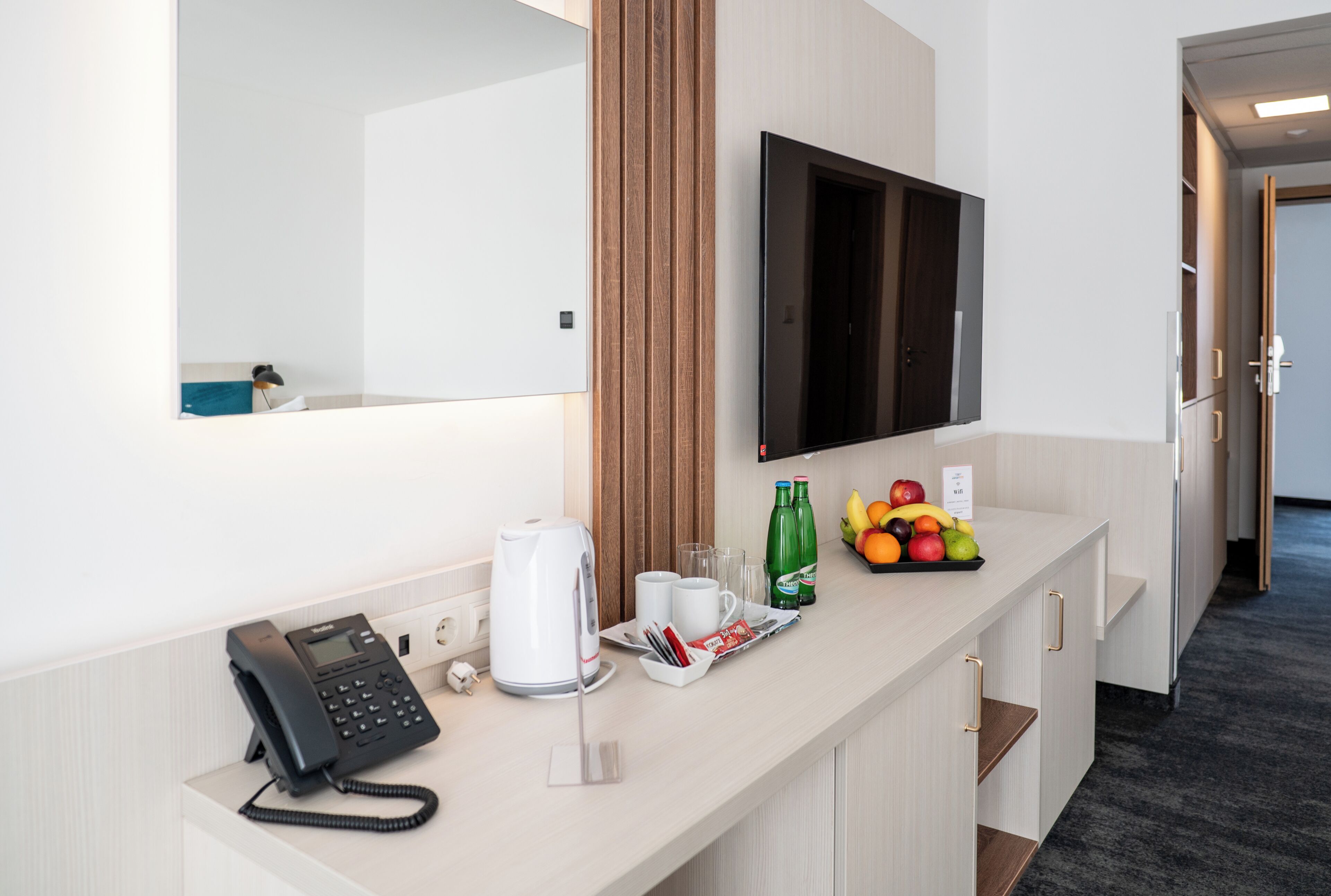 Et hotelværelse med skrivebord, telefon, fladskærms-tv, spejl og skål med frugt.