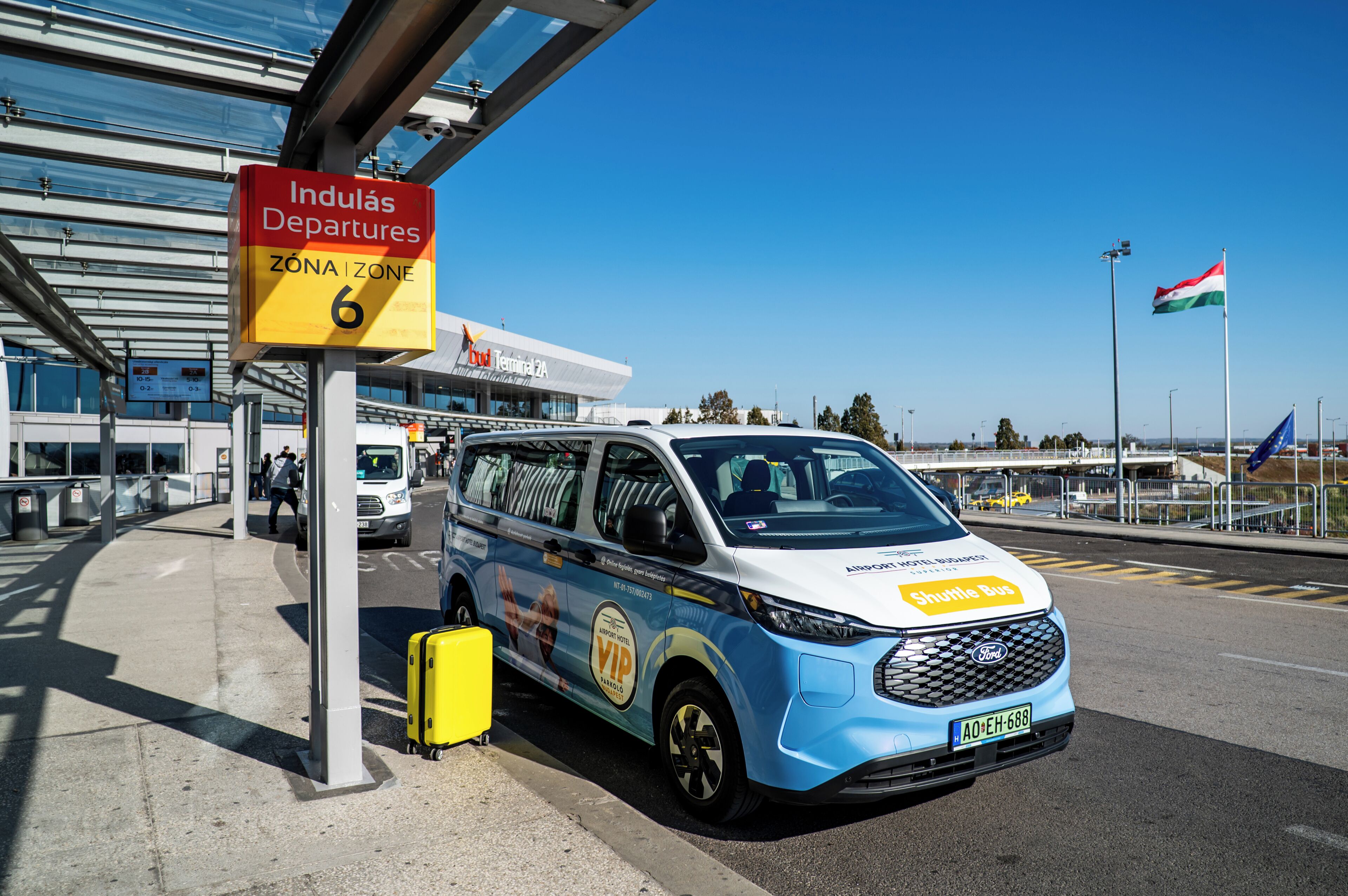 Un autobús lanzadera en un aeropuerto, con un cartel que indica 'Indulás, Zona de Salidas 6'.