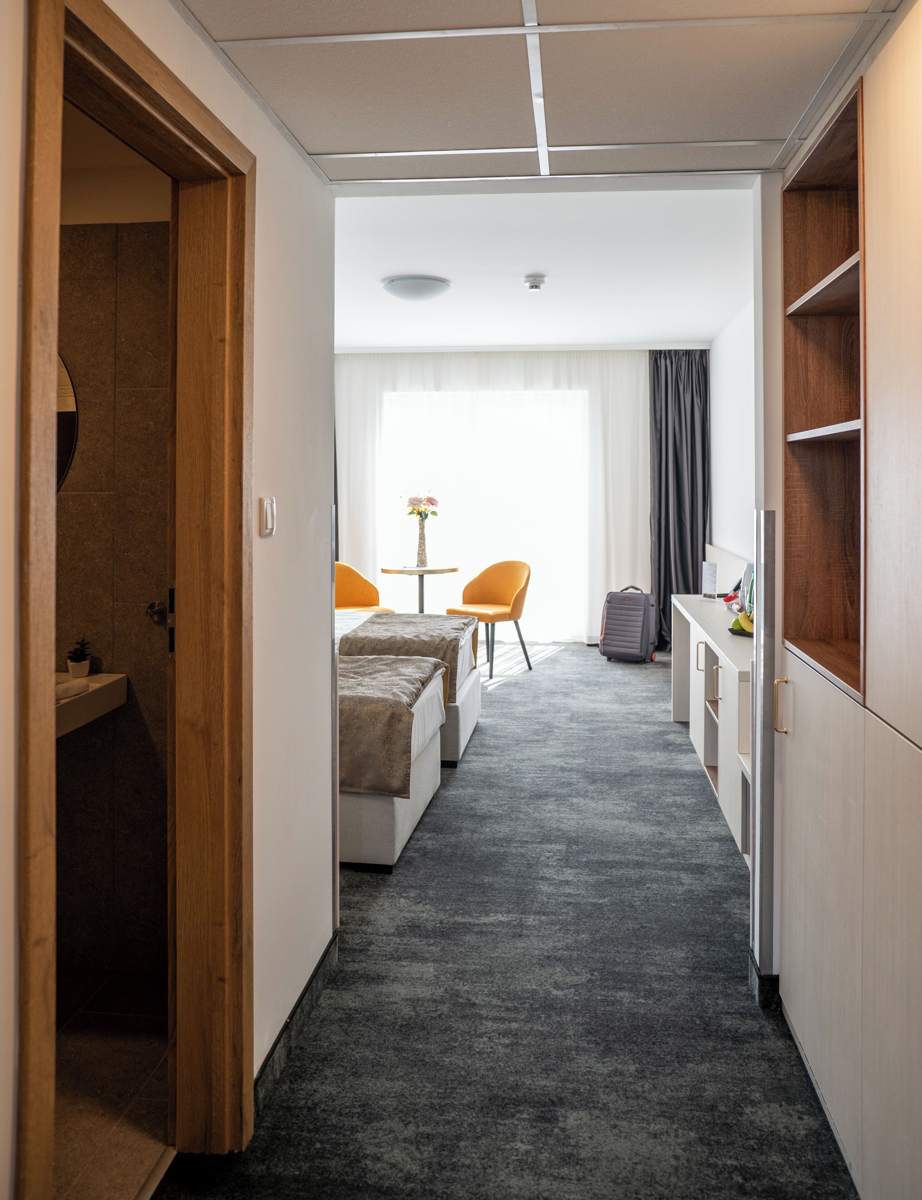 A hotel room with a bed, a desk, a chair, a small table with a vase, and a window with curtains.