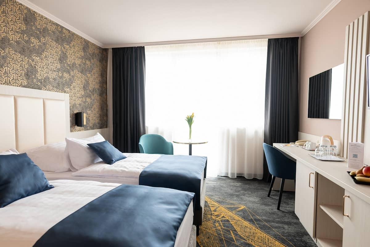 Habitación de hotel con dos camas, un escritorio, una silla, una mesita con un florero y un ventanal con cortinas.