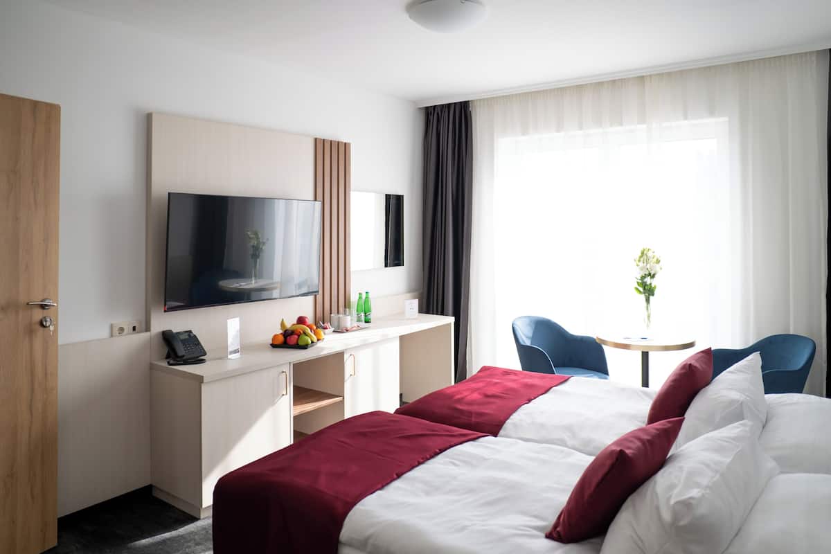 Habitación de hotel con cama, escritorio con televisor de pantalla plana, una silla y una pequeña mesa con un jarrón de flores.
