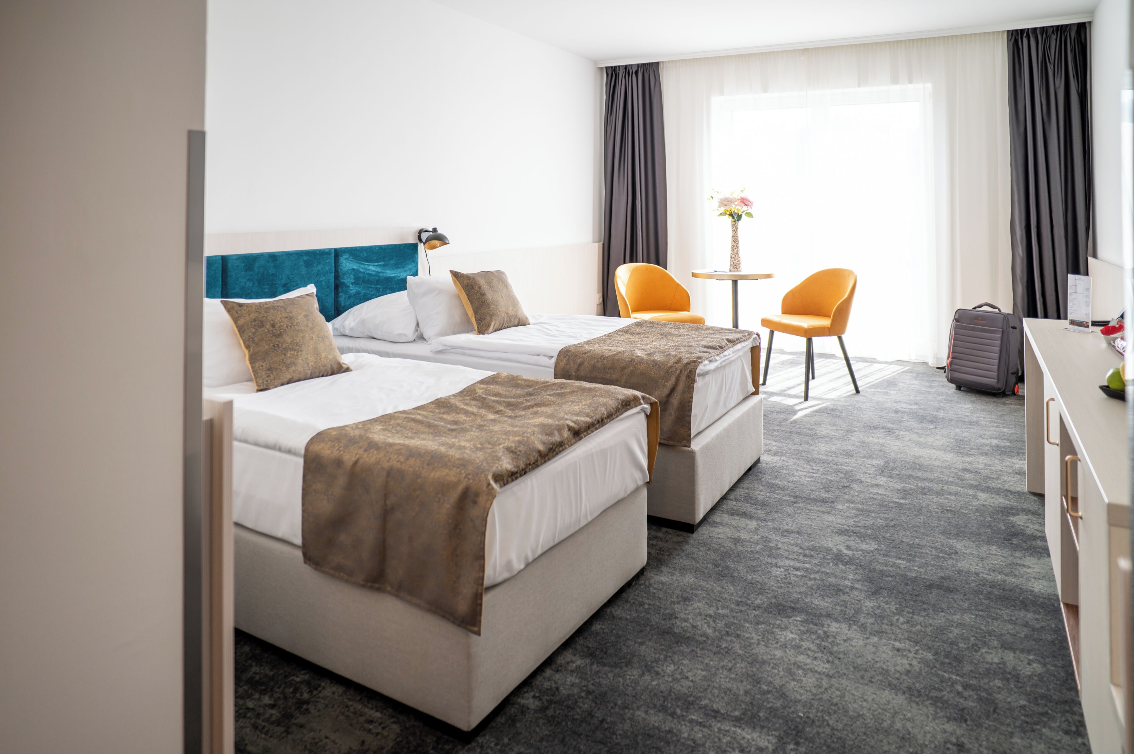 Quarto de hotel com duas camas, uma escrivaninha, uma cadeira e uma mesa pequena com um vaso.