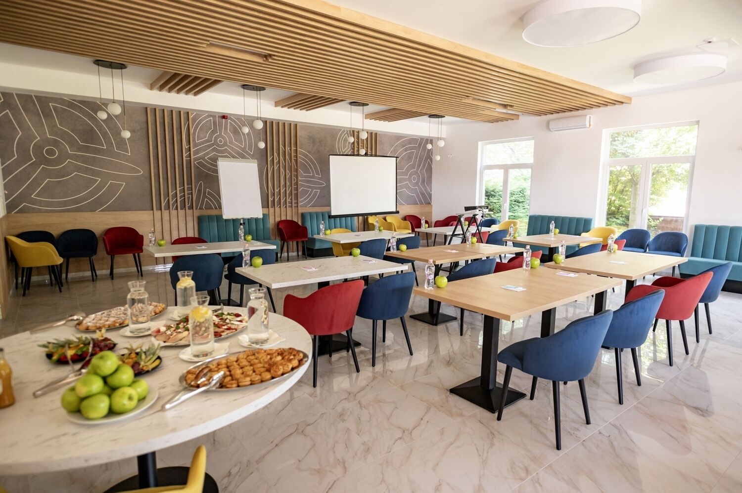 Un comedor moderno con varias mesas y sillas coloridas, una pizarra y un amplio ventanal que proporciona luz natural.