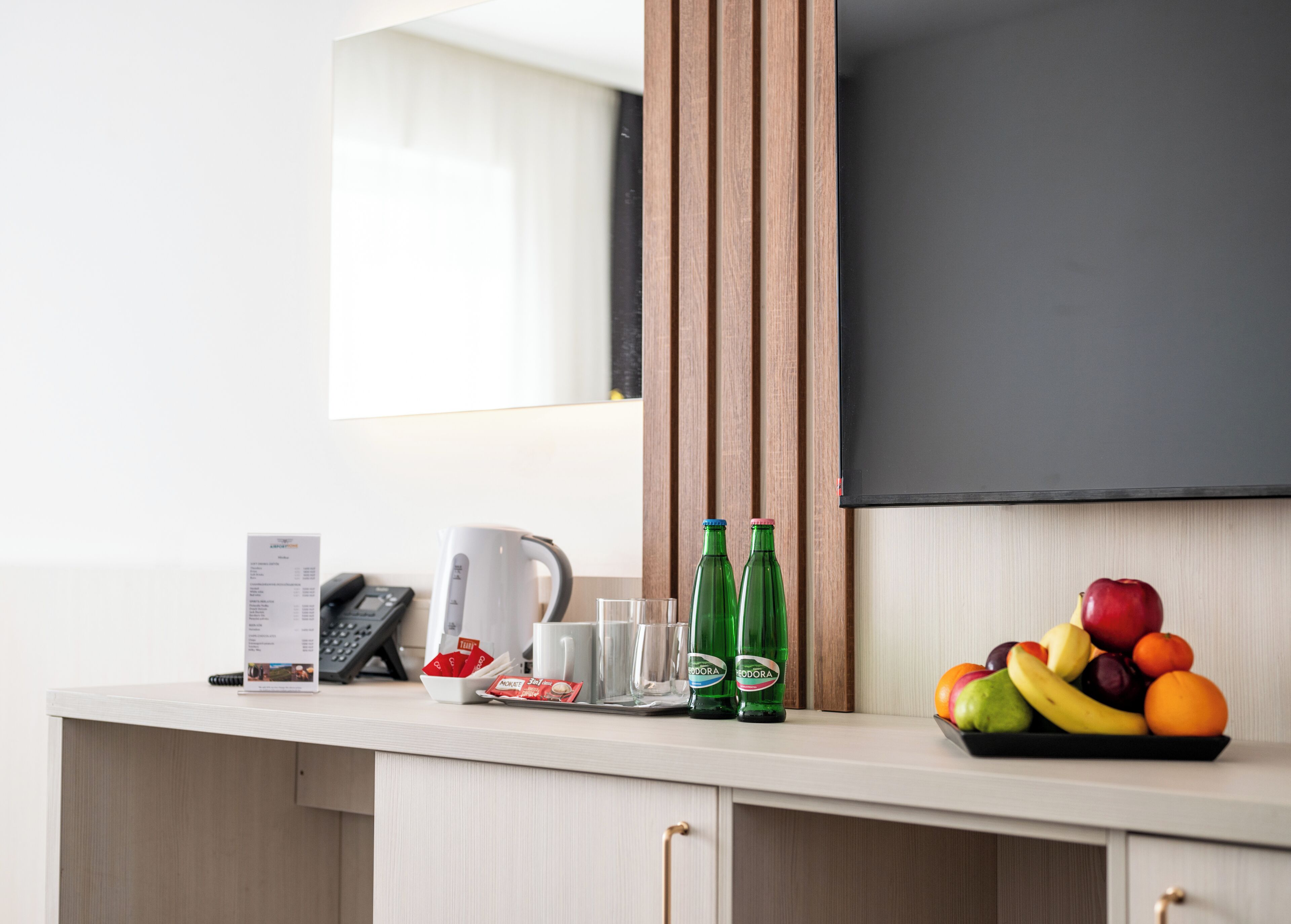 A kitchen counter with a kettle, a phone, a menu, two green bottles, and a fruit bowl.