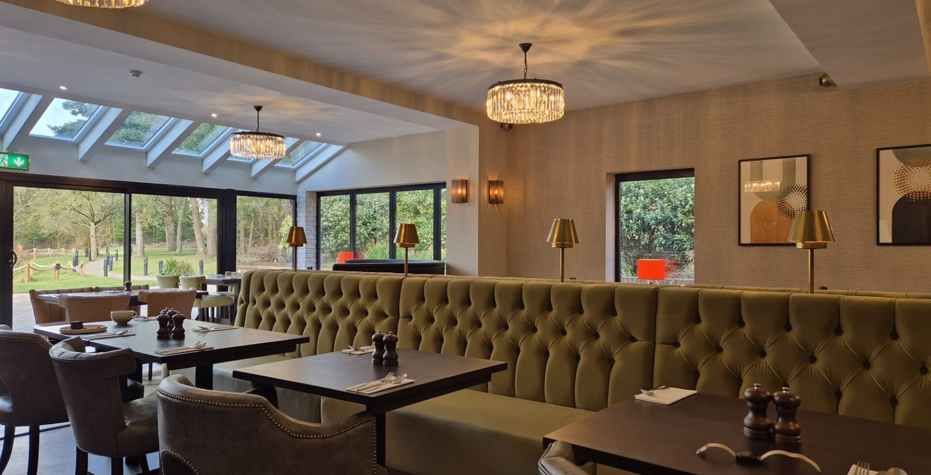 Un restaurante con una banqueta tapizada de verde, mesas y sillas. Hay ventanas amplias y un candelabro.