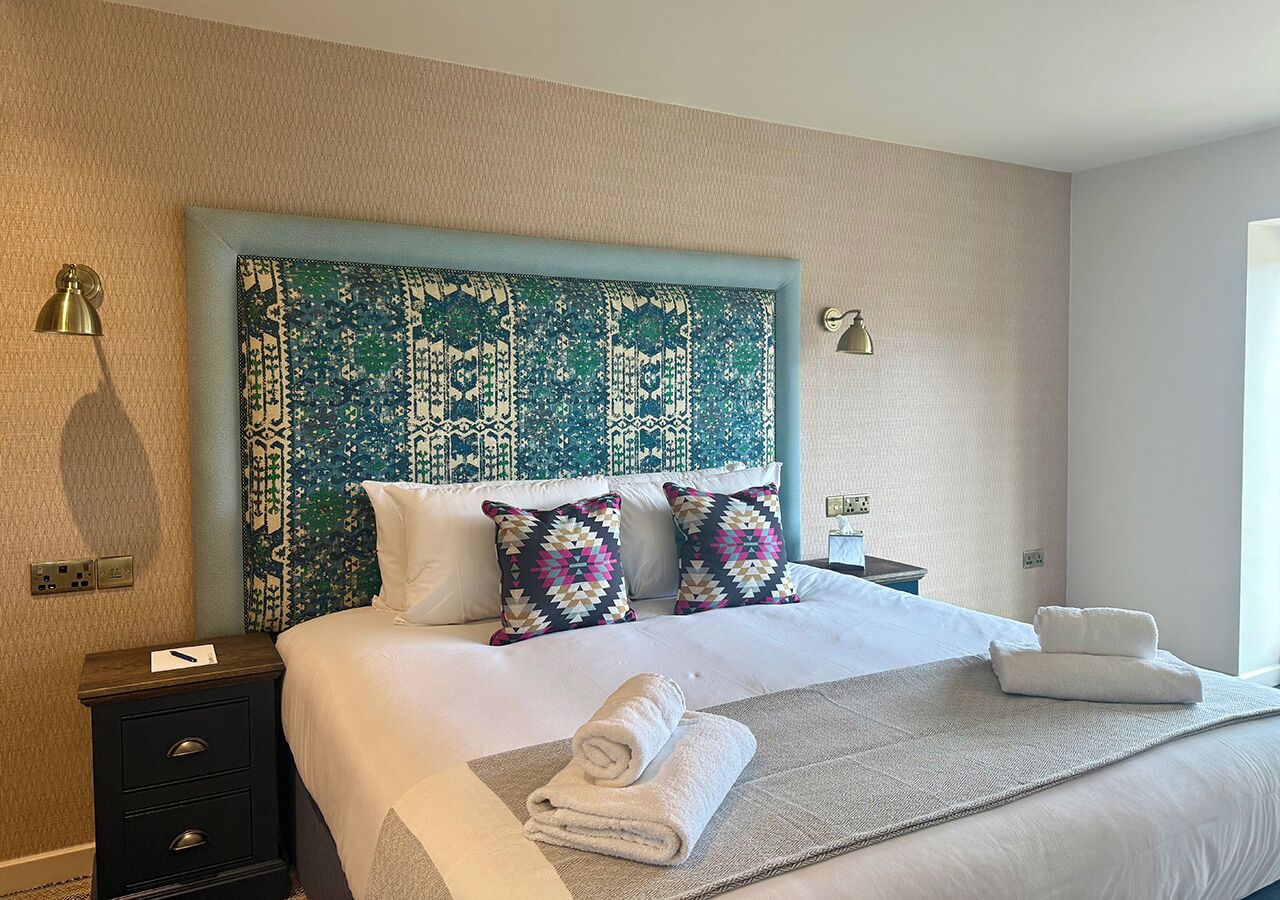 A hotel room with a large bed, a headboard with a patterned design, two bedside lamps, a nightstand, and a window.