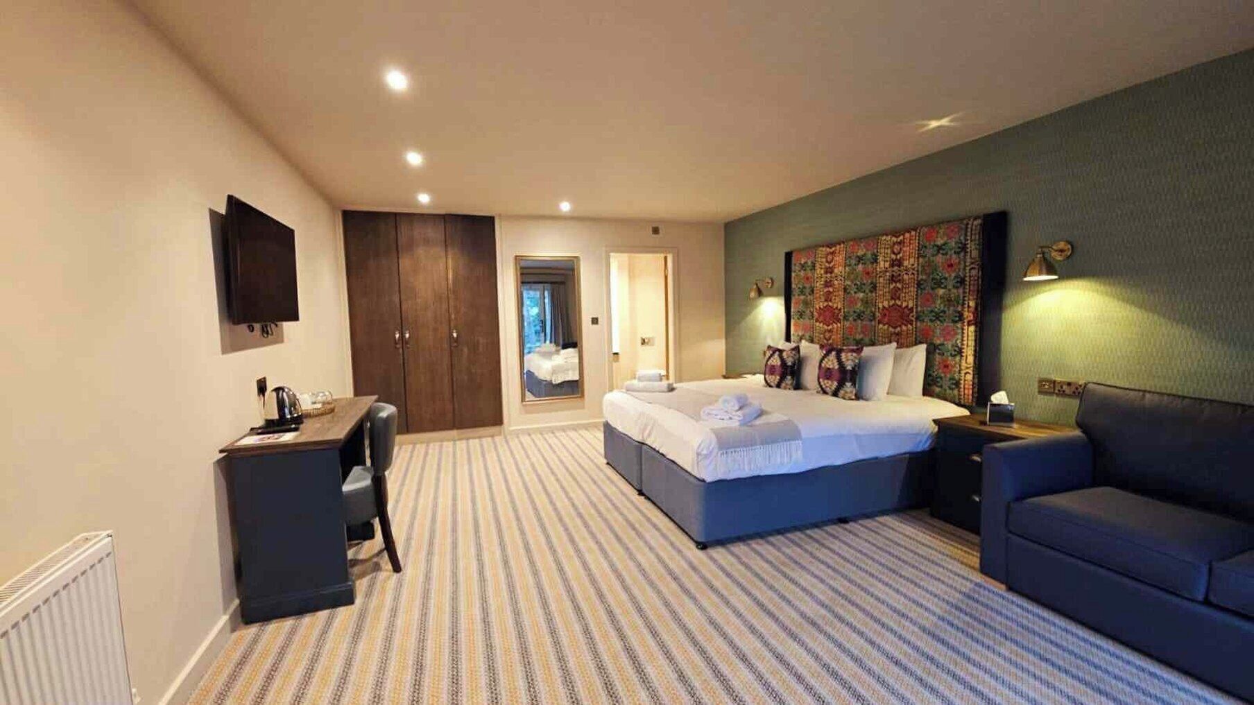 A hotel room with a large bed, a desk, a chair, a sofa, a TV, and a patterned carpet.