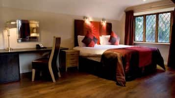 A bedroom with a wooden desk, a chair, a bed with red bedding, and a window with a view of trees.
