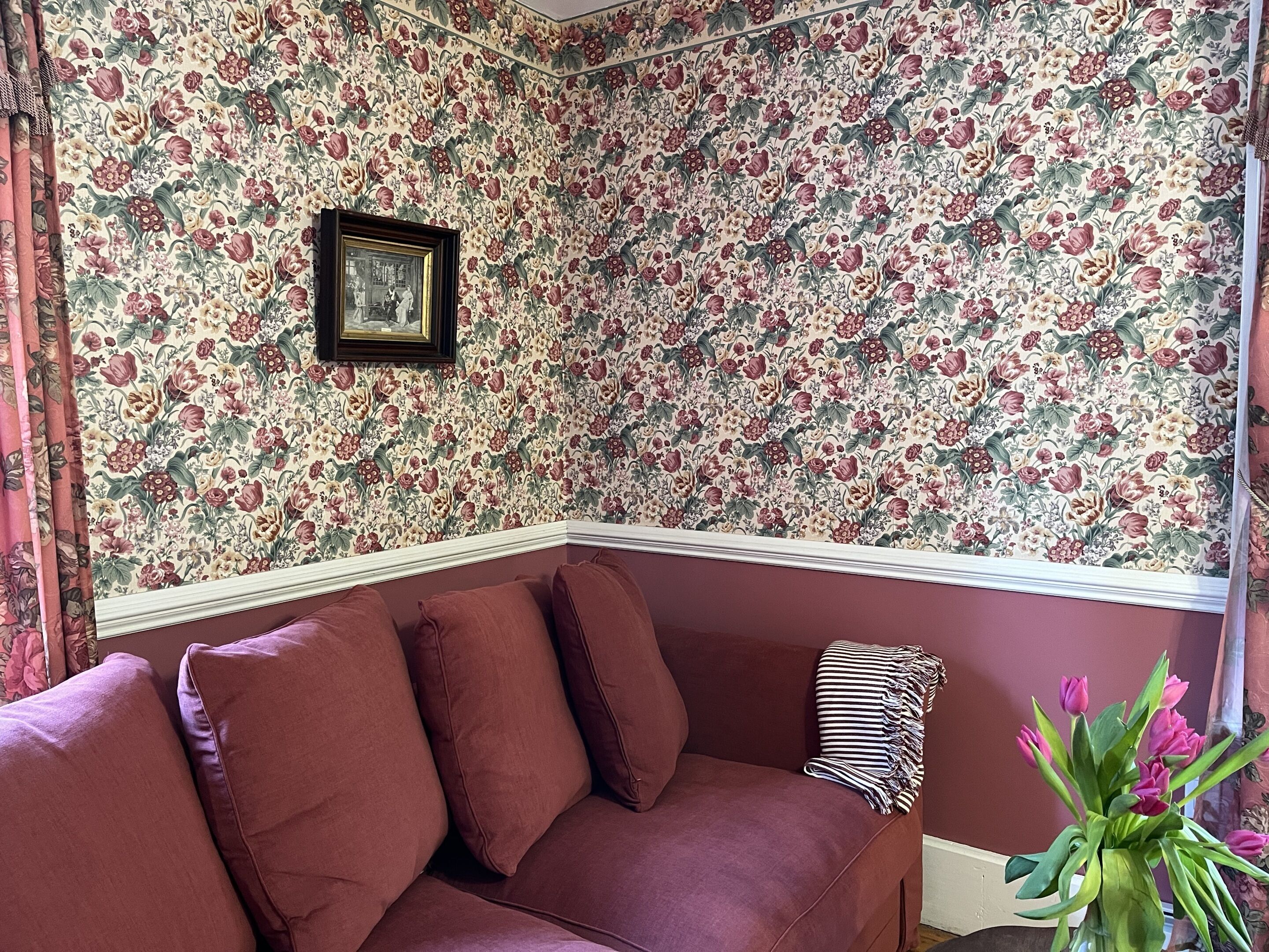 A corner room with floral wallpaper, a framed picture on the wall, and a purple sofa with pillows.