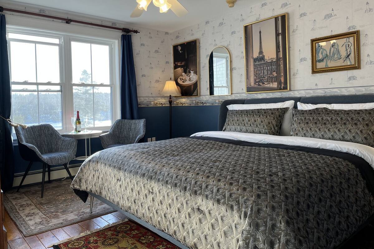 A bedroom with a large bed, a chair, a table with a bottle and glasses, a ceiling fan, and a window with curtains.