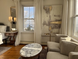 A hotel room with a bed, a marble-topped table, a chair, a large abstract painting, and a window with a view of trees and a building.
