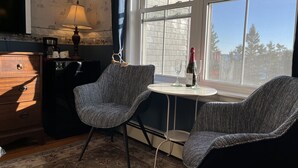 A cozy room with two patterned chairs, a small table with a bottle and glasses, a lamp, and a view of the outside through a large window.