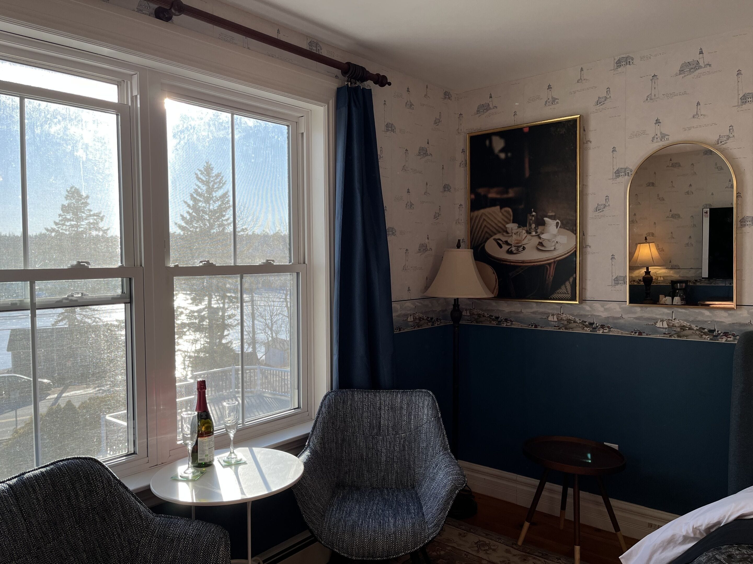 A room with a large window, a chair, a small table with a bottle and glasses, and a mirror on the wall.