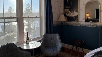 A room with a large window, a chair, a small table with a bottle and glasses, and a mirror on the wall.