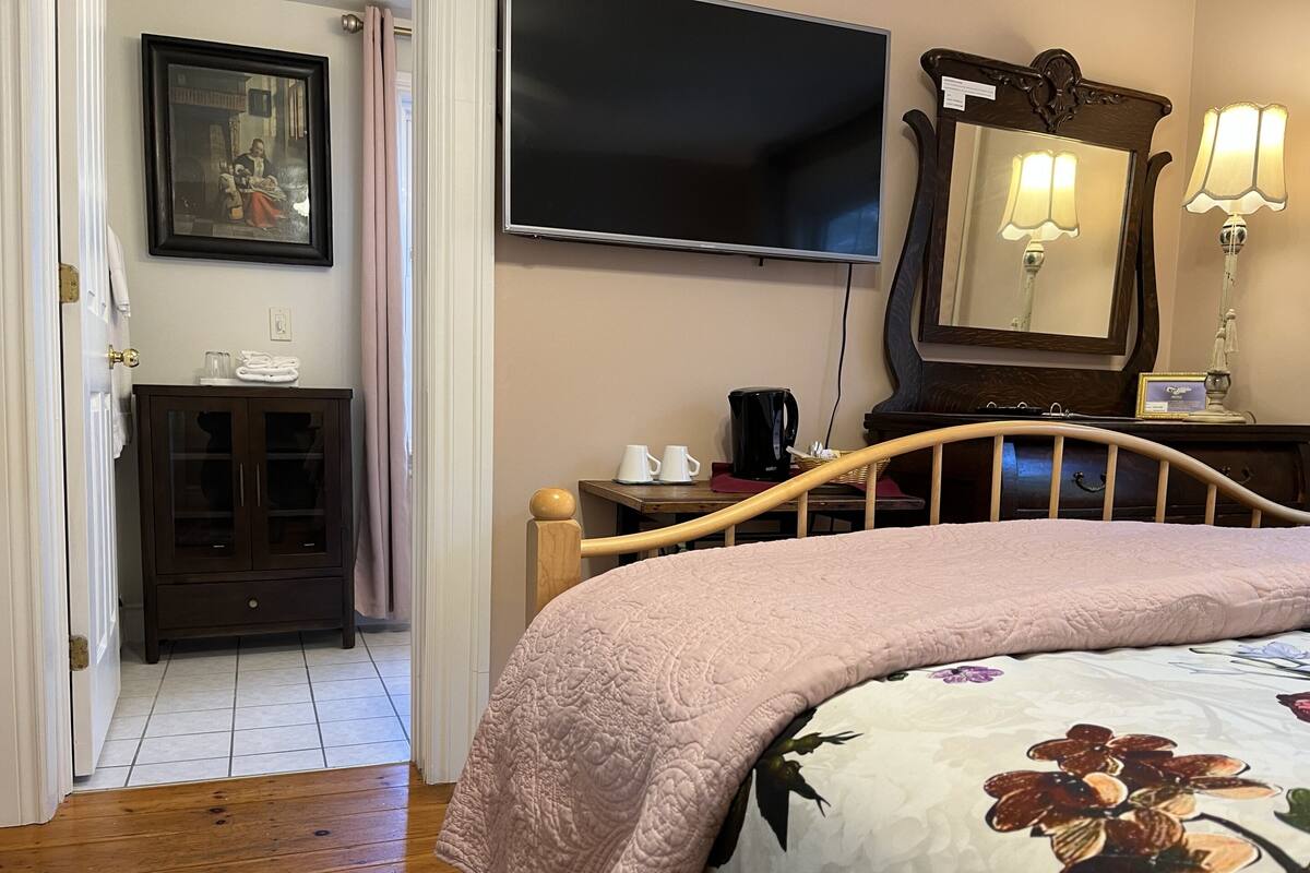 A bedroom with a bed, a television, a dresser, a mirror, and a door leading to another room.