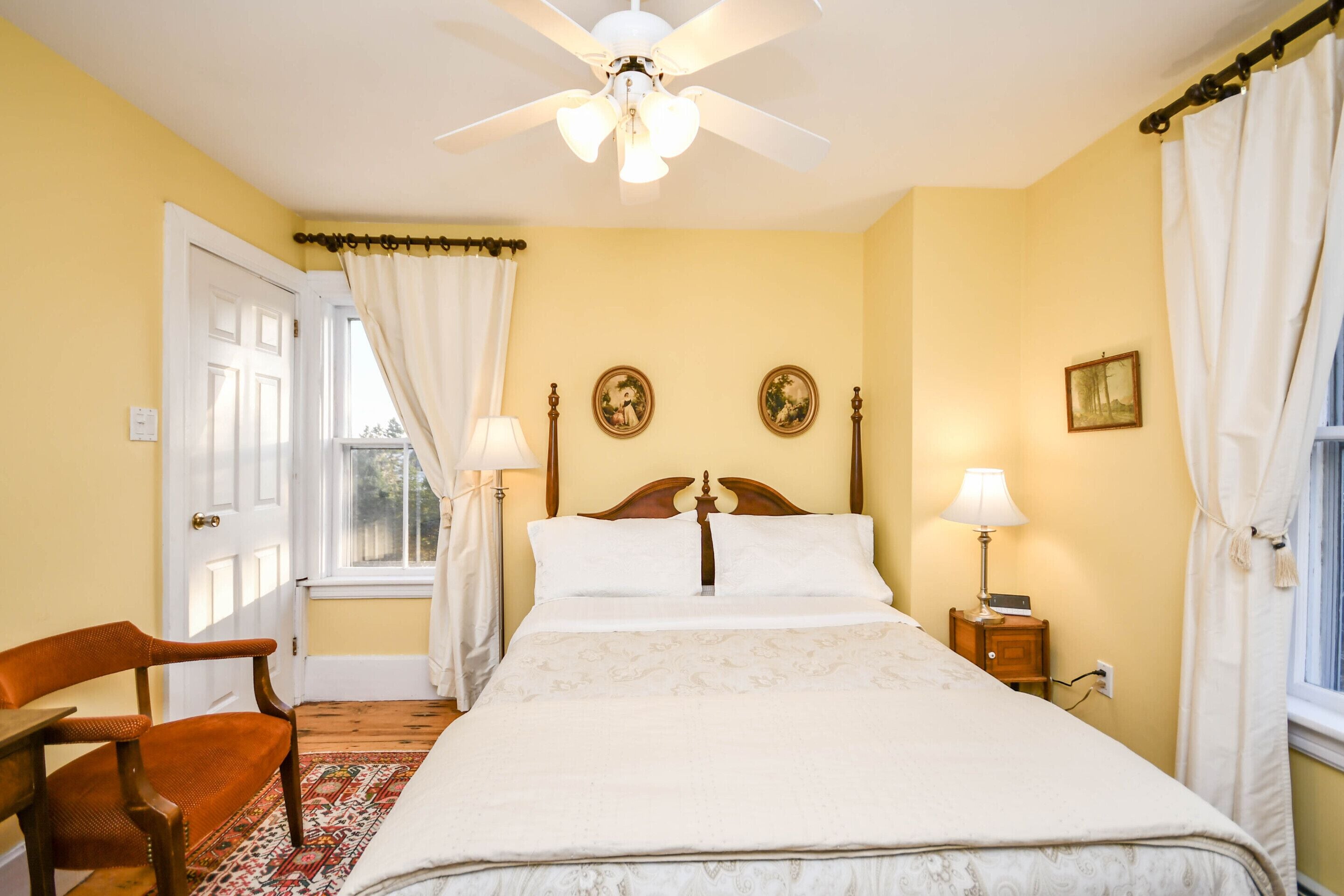 A bedroom with a bed, a chair, a nightstand, a ceiling fan, and two wall-mounted lamps.