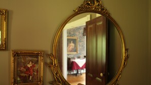 A room with a large ornate mirror, a framed painting, and a vase with red roses.