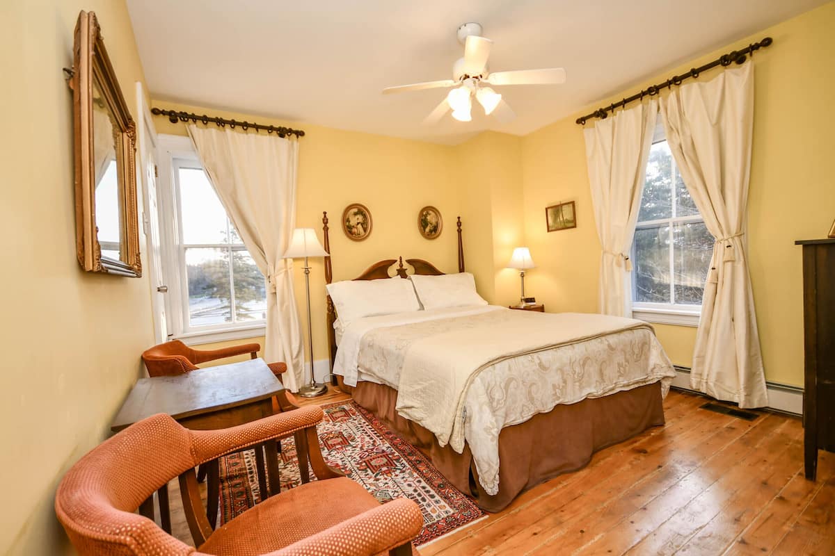 A bedroom with a bed, two bedside tables, a ceiling fan, a window with curtains, a wooden floor, and a chair.