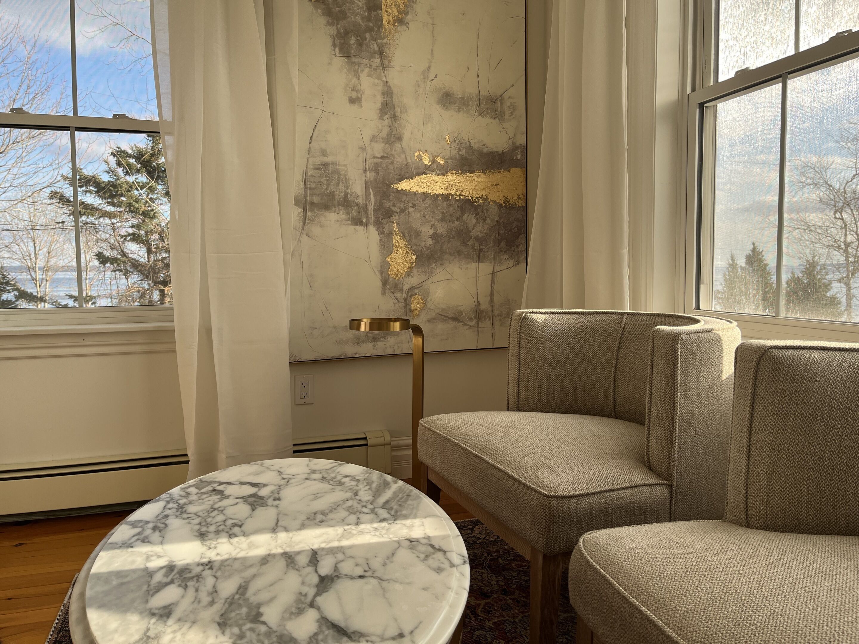 A modern living room with a marble table, a large abstract painting, and a view of trees through the window.