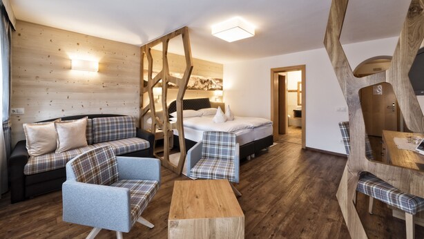 A cozy room with a wooden wall, a bed, a sofa, a chair, and a wooden table. - Hotel Interski (Santa Cristina Val Gardena)